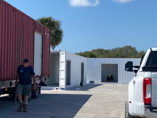 Man working near shipping containers and a white truck in a sunny outdoor setting.