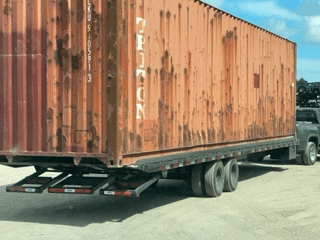 Rusty shipping container on trailer, parked on concrete. Blue sky background.
