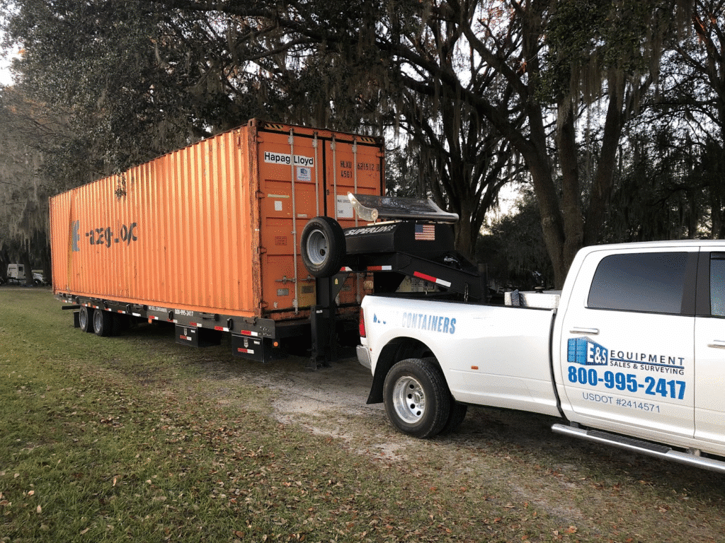 White truck towing orange shipping container in a forested area, highlighting logistics and equipment services.
