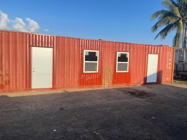 Red shipping container repurposed as an office, featuring white doors and windows, set against a palm tree backdrop.