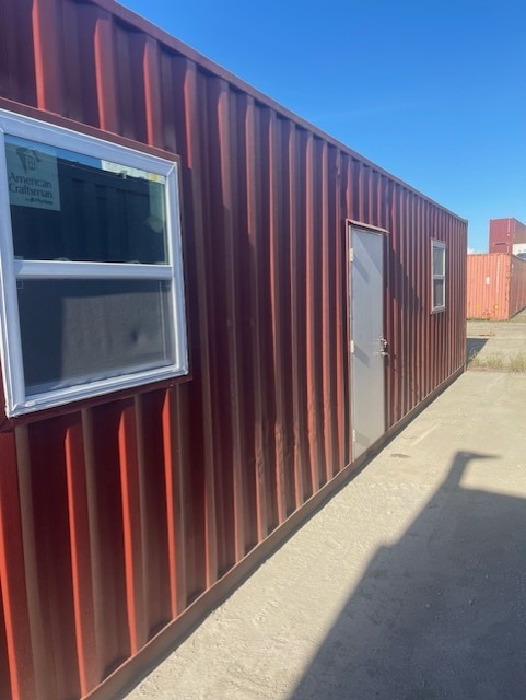 Red shipping container converted into an office with windows and door, placed outdoors under a clear blue sky.