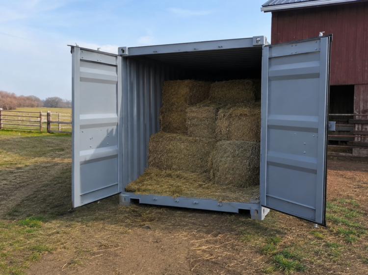 image A gray shipping container placed on a farm with its doors open, showing stacked square hay bales stored securely inside next to a traditional barn.