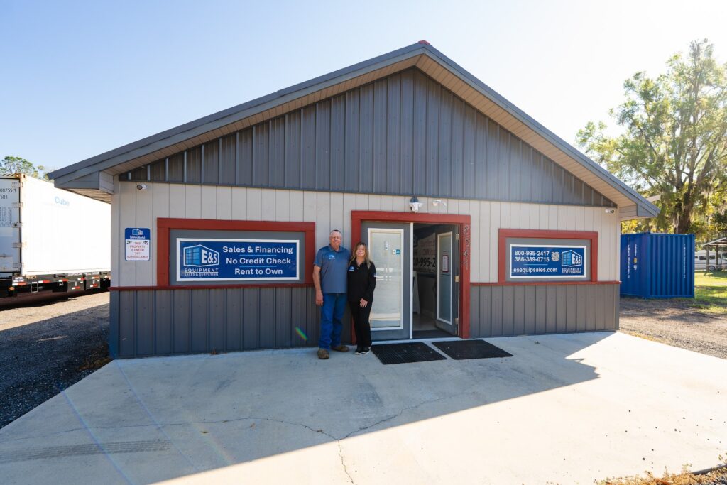 The owners of E&S Equipment Sales standing outside their local office, which features signs advertising their no-credit-check rent-to-own shipping container program.